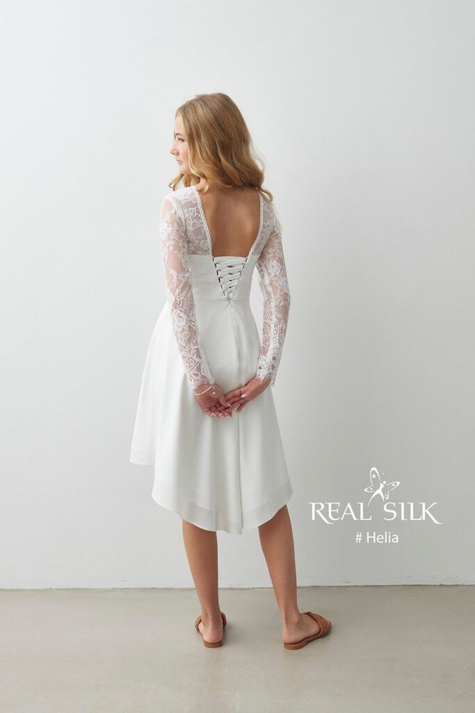 Real Silk Helia konfirmationskjole bagfra med dyb rygåbning, snørelukning, blondeærmer og high-low-skørt