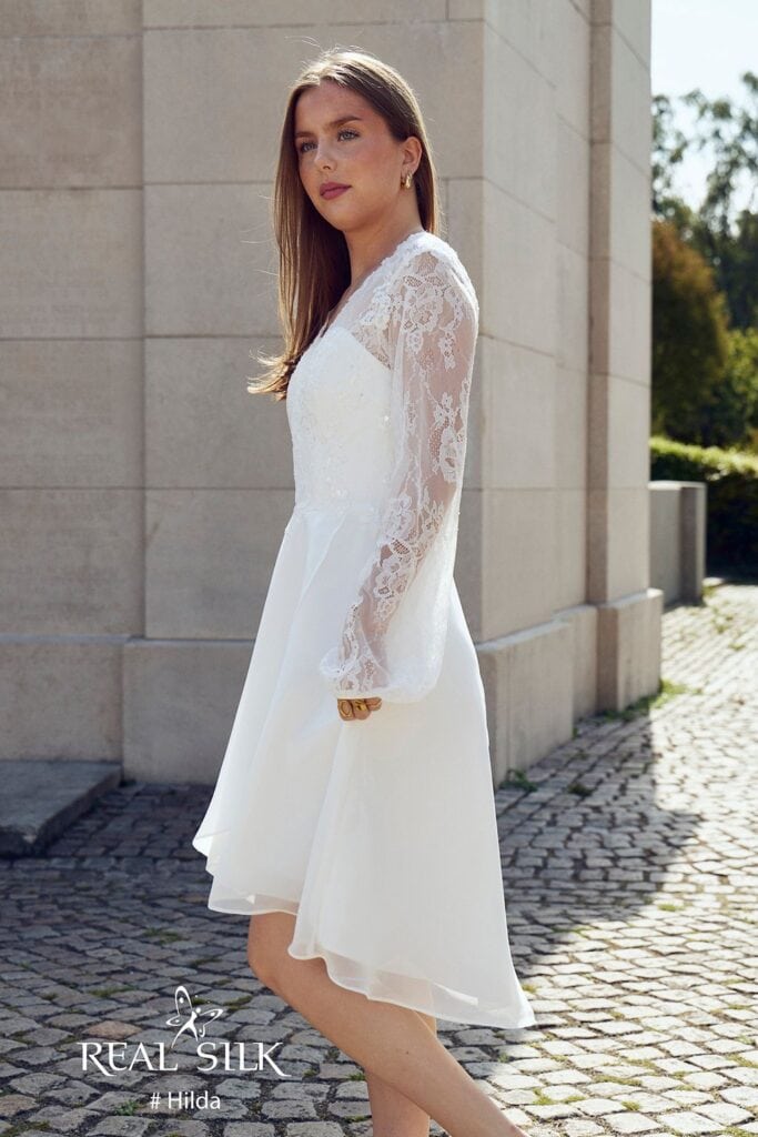 Real Silk Hilda konfirmationskjole fra siden med transparent blondeærme og high-low-skørt med længere bagstykke